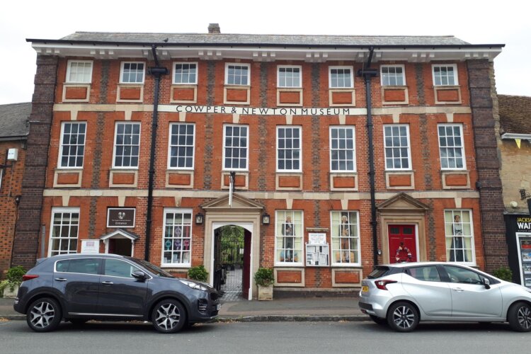 A large red brick building with many windows. Enter via the shop. There is a sign at the top of the building saying 'Cowper & Newton Museum'