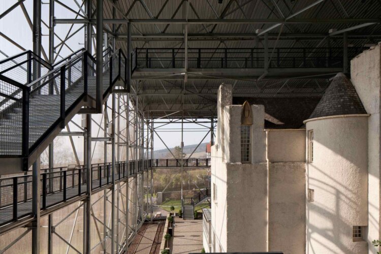 A white, two‑story building with a steep dark roof inside a large metal-and-glass structure.