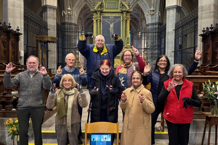 The smiling members of Heart & Lungs Singing Group, looking at the camera and crossing their fingers communicating that we are funded by the National Lottery Community Fund.