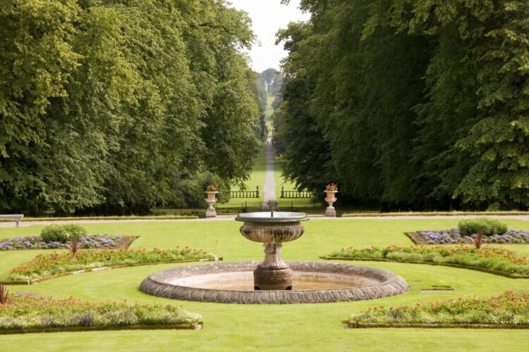 A round stone fountain in a formal garden with a long, tree‑lined path behind it.