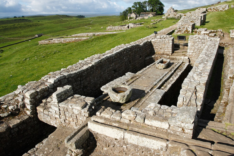 Ancient Roman stone ruins with an open rectangular structure on grassy hills under a blue sky. The scene conveys a sense of historical tranquility.