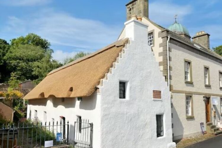 A whitewashed cottage with a thatched roof beside a larger stone building, set along a quiet road under a bright blue sky.