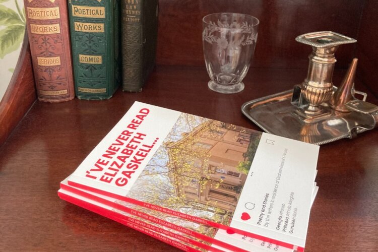 Pile of new red and white books on a wooden Victorian bedside table. There is a silver candlestick holder, water glass and old books in the background.