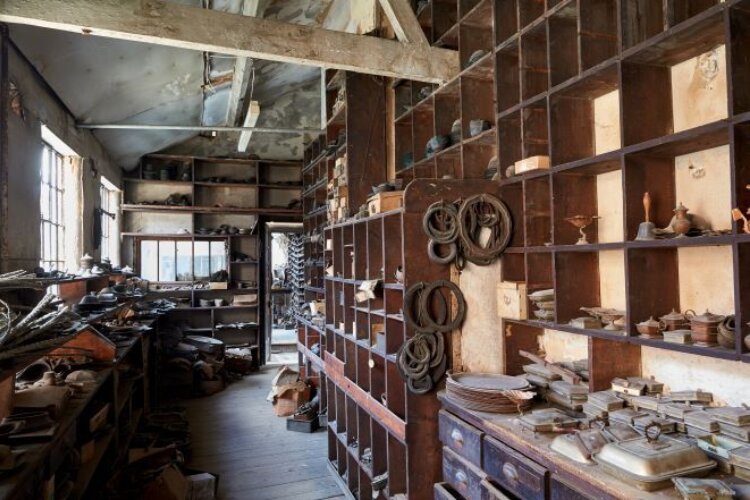 A cluttered workshop with wooden shelves filled with various tools and equipment. Coiled ropes hang on the wall, and natural light streams in through large windows, creating a nostalgic and rustic atmosphere.