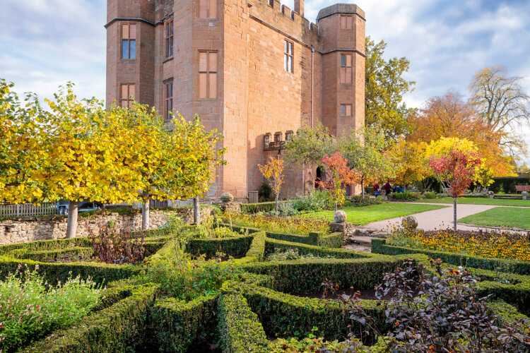 A historic stone castle with tall towers is surrounded by lush, geometric hedges and colourful autumn trees, creating a serene and majestic landscape.
