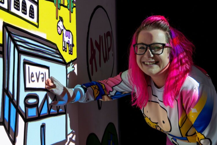 A person with long pink hair smiles at the camera. They stand next to a wall with colourful doodle style projections on it.