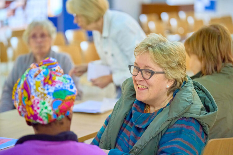 A lady with glasses laughing with another person with a colourful hat on (face not shown) at the cafe