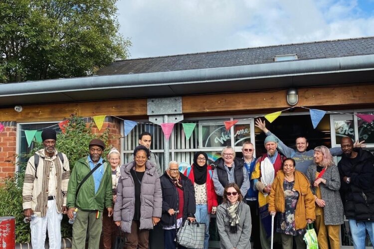 “A diverse group of adults standing and smiling outside a community building decorated with colourful bunting, posing together near the entrance on a paved area.”