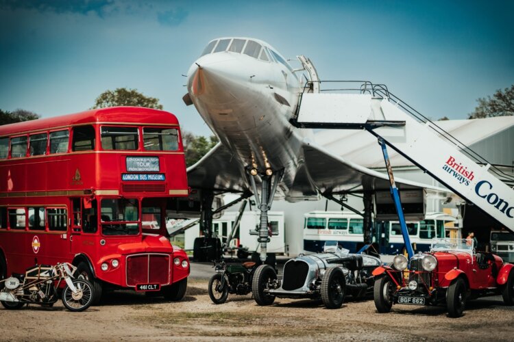 A classic red double‑decker bus from the London Bus Museum collection, several vintage racing cars, and an old motorcycle from the Brooklands Museum Collection are displayed in front of a British Airways Concorde. Other historic buses and vehicles are visible in the background at Brooklands Museum.