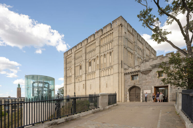 A historic stone castle with tall rectangular walls and narrow vertical windows, viewed from a pathway leading to its entrance. The castle has a fortified gateway with a large wooden door and a few visitors standing near the entrance. To the left, a modern cylindrical glass structure contrasts with the medieval architecture. A black metal railing runs along the pathway, and a tree with green leaves frames the right side of the image. The sky is bright blue with scattered white clouds.