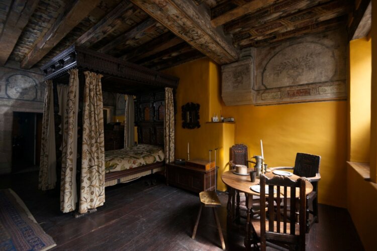 A historic room with blue walls, wooden beams, a large storage cabinet with textiles, a wooden worktable, and two curtained windows lighting a small table and chairs.