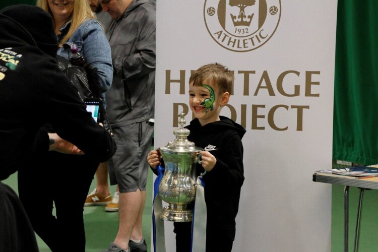 A young boy grins at an adult taking a photo, while holding the FA Cup. Behind him is the Wigan Athletic Heritage Project banner.