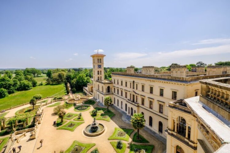 A grand historic building with a tall, square tower overlooks a manicured garden featuring a central fountain, surrounded by lush green fields under a clear blue sky.