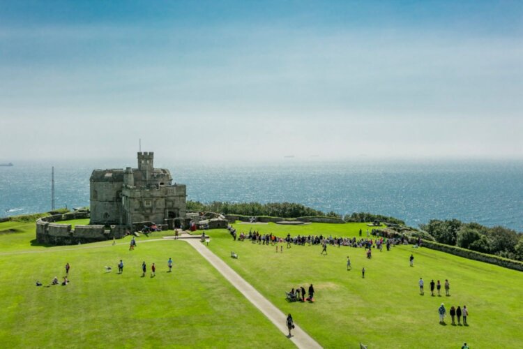 A historic stone castle stands on verdant cliffs overlooking a vast ocean under a clear blue sky. Visitors wander the lush green lawn, evoking a serene, leisurely atmosphere.