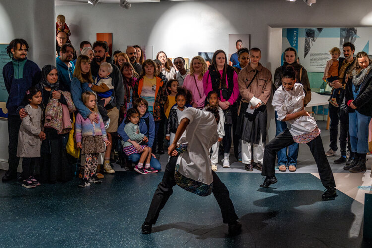 Two dancers perform in a museum gallery watched by a standing crowd of adults and families