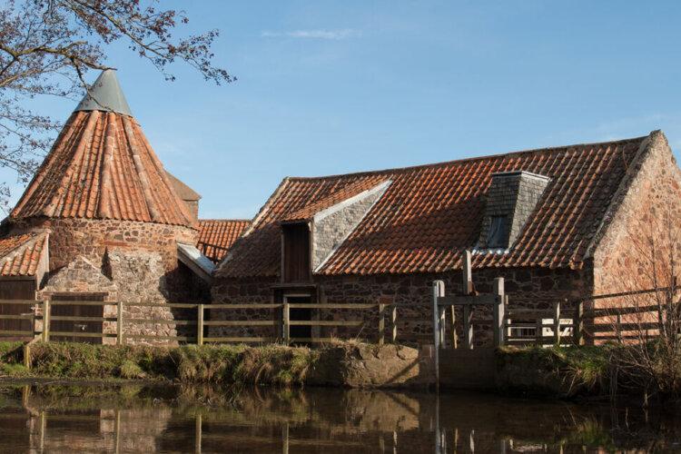 A historic brick mill with red‑tiled roofs, including a tall conical mill structure, beside a calm waterway under a clear blue sky.
