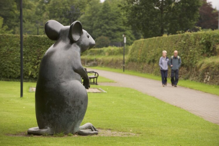 A large statue of a mouse sits in a grassy park area beside a path, while two people walk along the path in the background.
