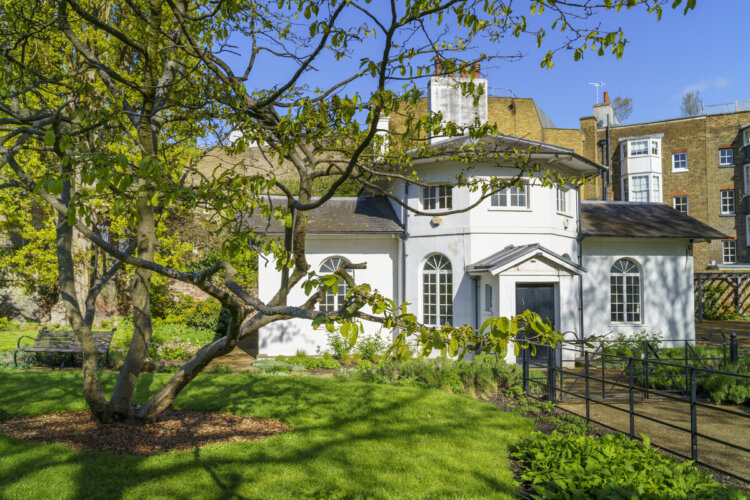 The image shows a Victorian white cottage surrounded by gardens, with a tree in the foreground.