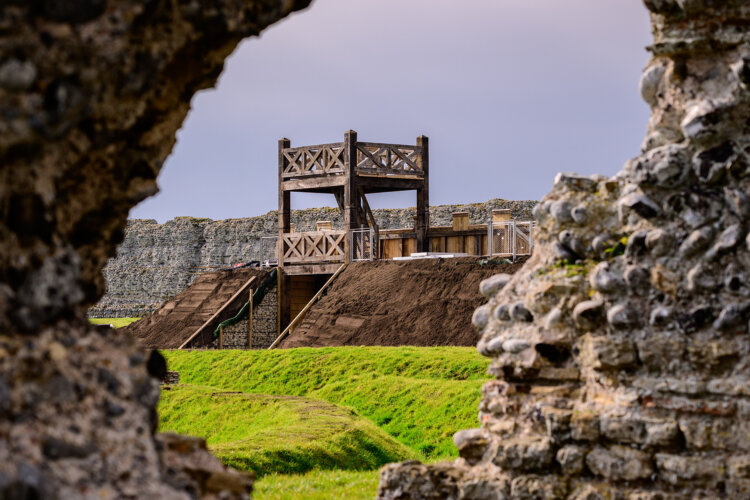 View of a wooden fort structure through a stone-arch ruin, with a lush green grass foreground and an overcast sky, conveying a sense of historical intrigue.