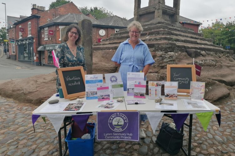 Two smiling ladies stand behind a table with information and signs that read 'volunteers needed' for The Sanctuary Hub. They are in a village/town location with a monument behind them.