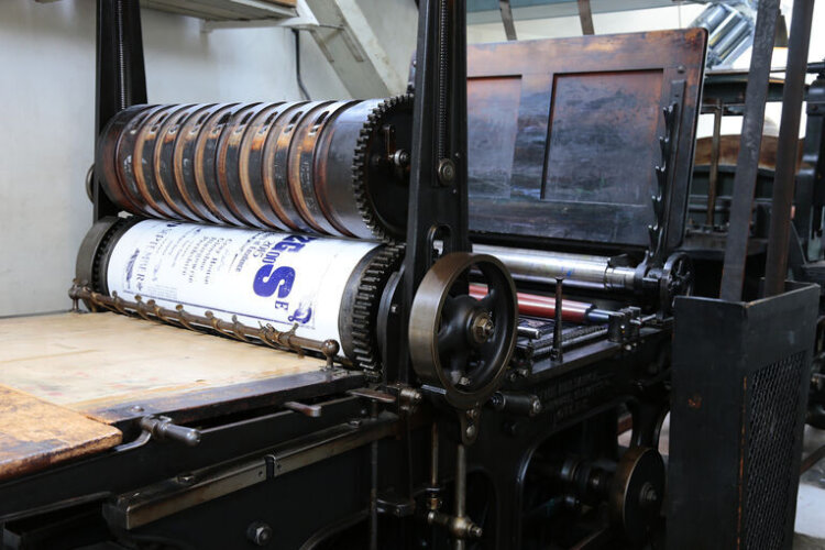 An old industrial printing press with large rotating rollers, gears, and metal components, holding a sheet of printed material between the cylinders.