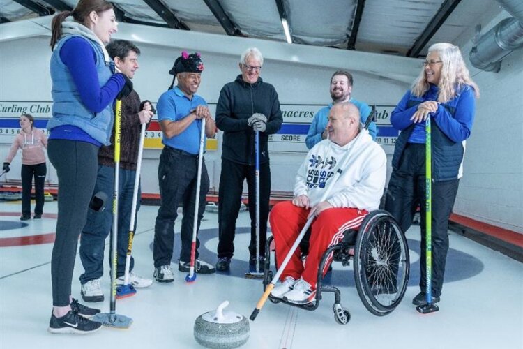 A group of adults stand on an indoor curling rink, smiling and talking while holding curling brushes. One person in a wheelchair prepares to push a curling stone on the ice as the others watch.
