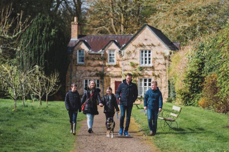 Three children, two adults and a dog walk down a path away from an old house