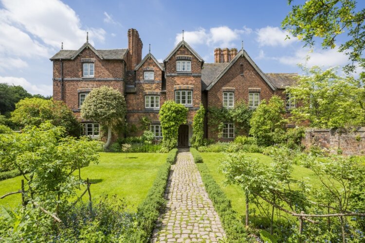 A large brick house stands behind a luscious green garden