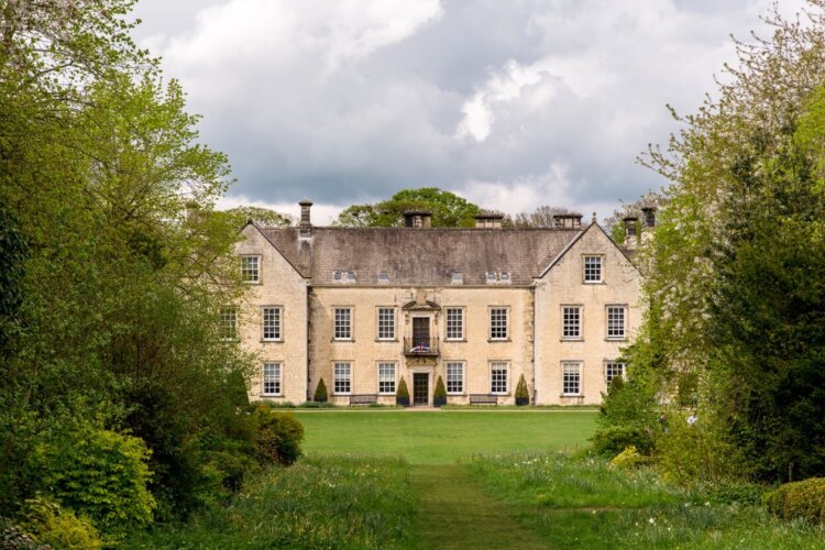 A large cream manor house with a mature green garden in front of it
