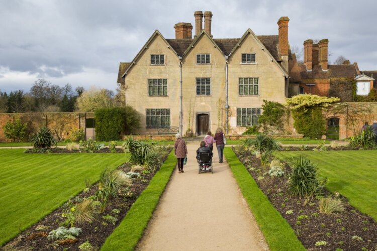 Four people, including one in a wheelchair, are walking down a path with gardens either side and a big cream coloured house at the end