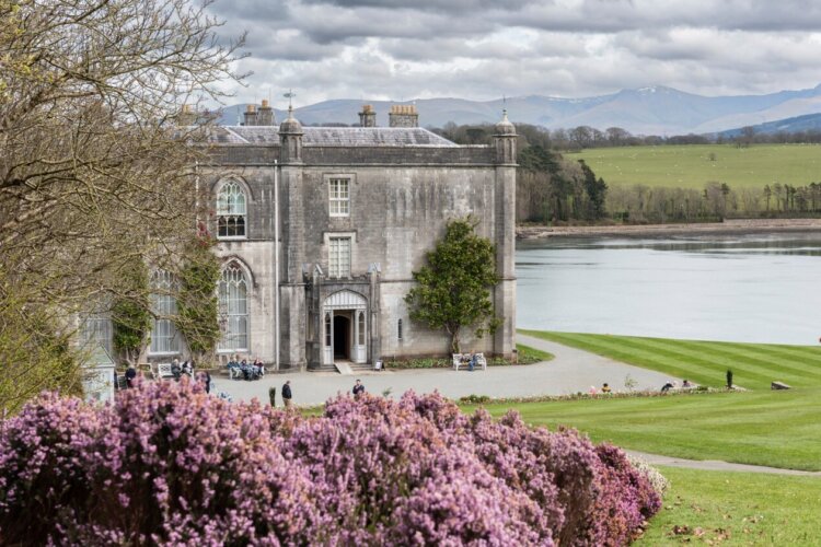 A large grey house with mature gardens next to a body of water