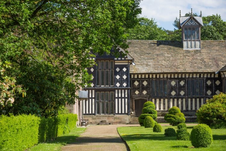 An old tutor building with black panelling sits behind a big tree and well manicured garden