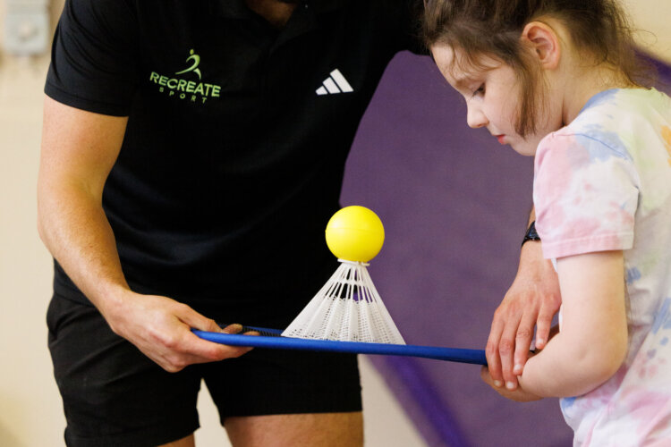 A man with brown hair and wearing a black t-shirt, stands next to a child with brown hair. The child is holding a badminton racket with a large shuttlecock balanced on top.