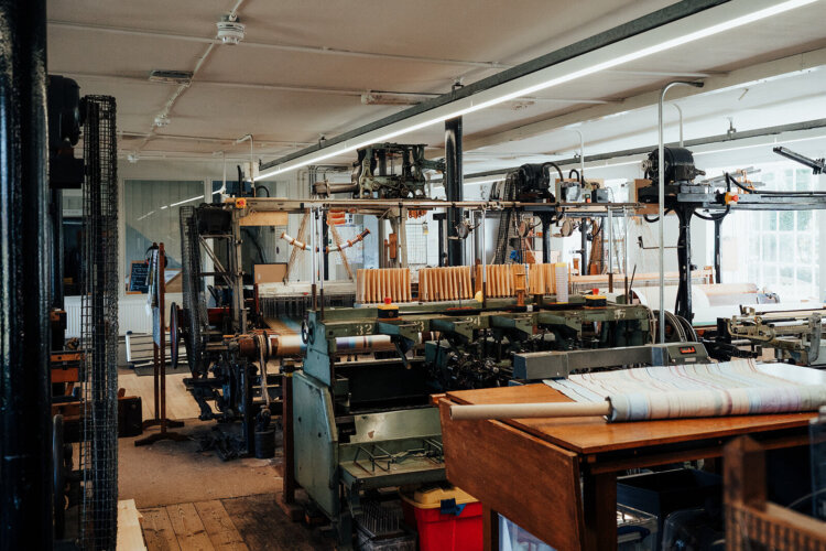 Historic Victorian looms inside Whitchurch Silk Mill, showcasing rows of black metal weaving machines with wooden shuttles and threads in place, set in a bright workshop featuring wooden floors and large windows that let in natural light.