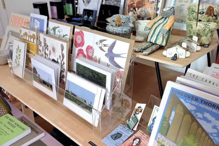 Image is of a table and shelf, showing a mix of art-based items and gifts, including small ceramic sculptures, gift cards, books and candles.