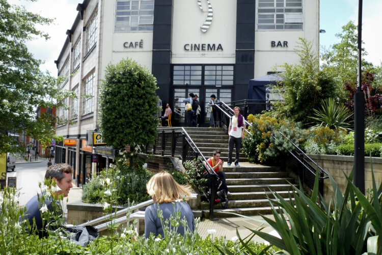 A black and white building sits behind some stairs leading down to a green area with people seated on both the stairs and benches