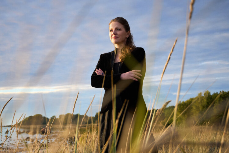 A white woman wearing all black looks into the distance, arms folded. Background in a verdant coastline and the foreground features long, marram grass.