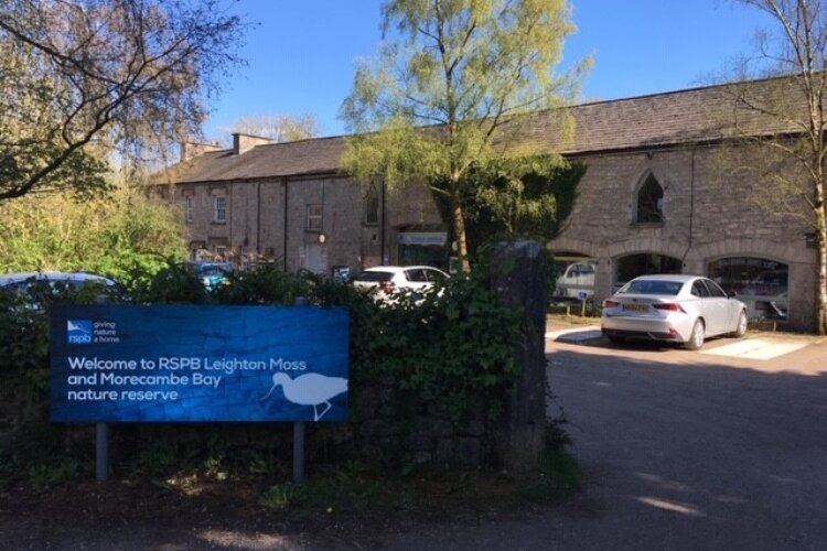 Alt=“The entrance to RSPB Leighton Moss reserve and visitor centre, with a sign in the foreground and buildings in the background”