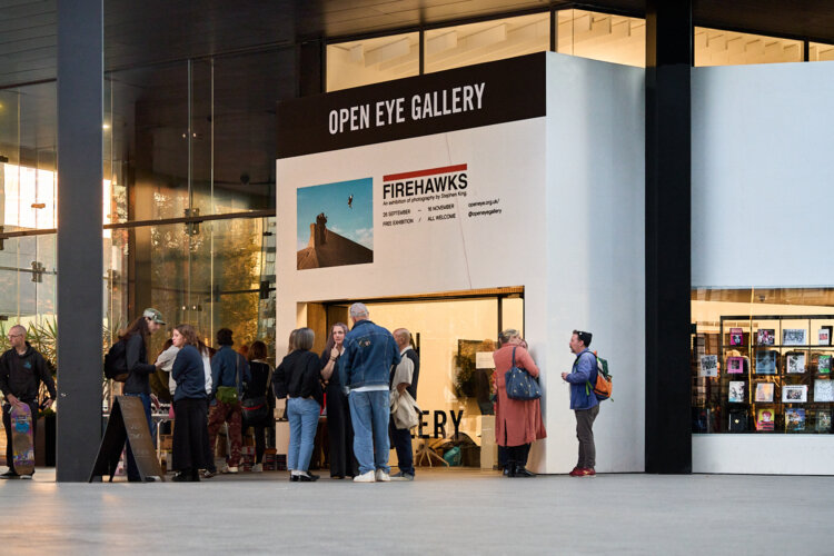 People are standing in front of the Open Eye Gallery in a covered atrium space during the launch event. The gallery is a small white buildings with its name above the door. There are some photos on the outside walls of the gallery.