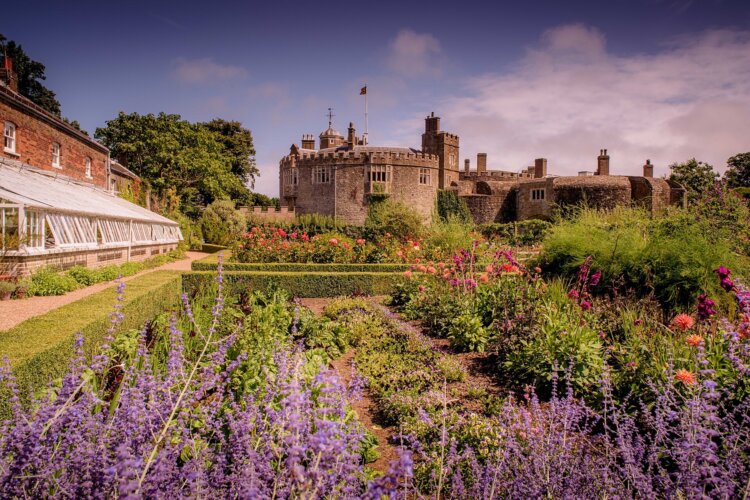 A historic stone castle with a central tower stands under a partly cloudy sky. In the foreground, a vibrant garden blooms with colourful flowers and lush greenery.