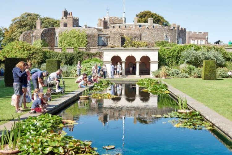 People gather around a reflective pond in a lush garden, overlooked by an old stone castle. The scene is lively and relaxed, on a clear day.