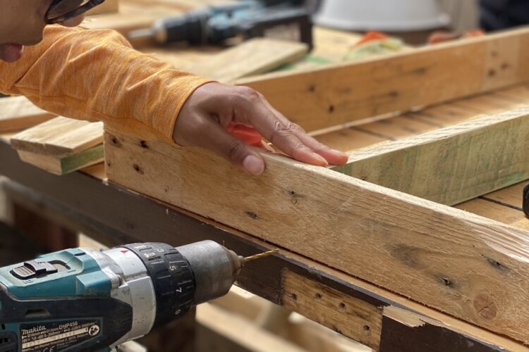 A person wearing a headscarf and glasses using a power drill on a timber frame