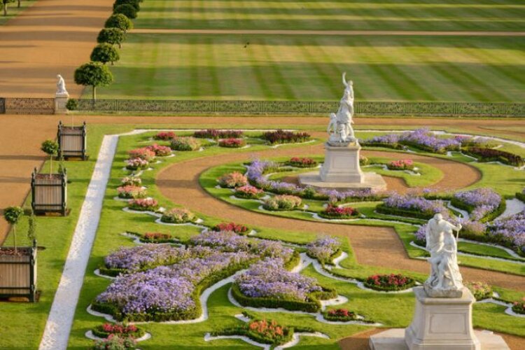 Elegant garden scene with colourful flowerbeds in swirling patterns, bordered by neat hedges. Marble statues stand prominently on pedestals, evoking a tranquil, classical ambiance.