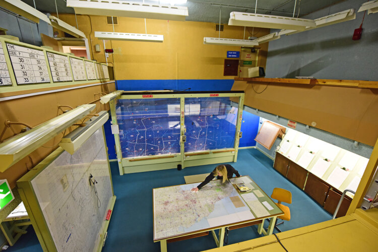 A person examines a large map on a table in a control room with blue walls and maps on display. The setting is strategic and focused.