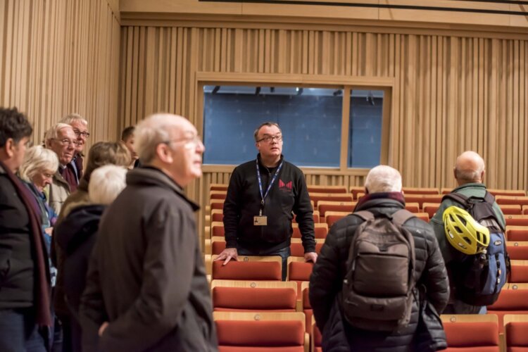 A photo of a man talking to a small group of people in the seating are of a music venue.