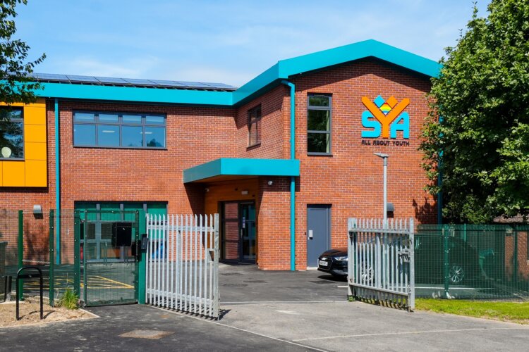 A red brick building with a blue roof and guttering (with a little yellow section around the side window) has the letters S Y A on the front. The gates are grey with green fencing and a blue car parked out front.