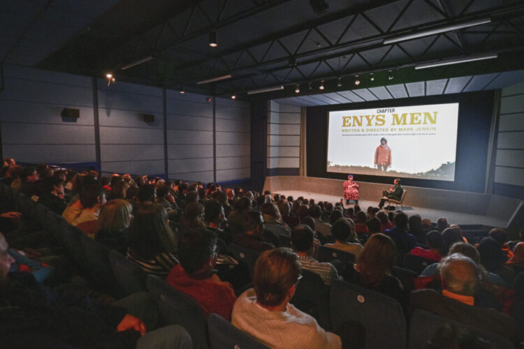 A  view of a sold out screening in Cinema 1 in Chapter with the screen on and the stage set for a Q&A with two figures seated on the stage with a table between them. The room is has blue panels and blue seats.