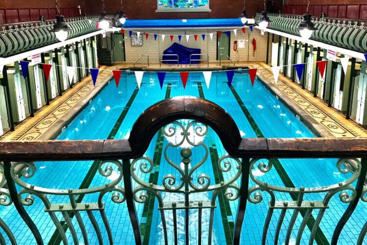 Image of bright blue Edwardian style swimming pool, large stained glass window at the end of pool hall. Ornate wood balcony in foreground and large roof lanterns.