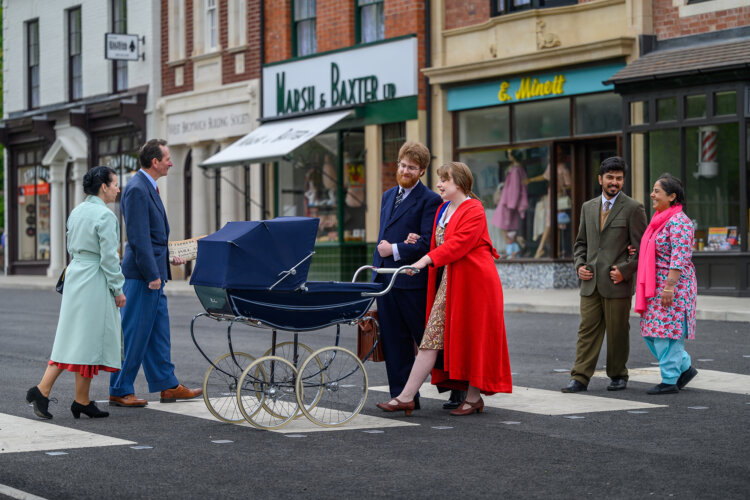 A group of six historic characters dressed in 1950s clothing is crossing over a zebra crossing on the Museum's 1940s-60s high street. There are shops in the background including a record shop, butchers, ladieswear shop and building society.
