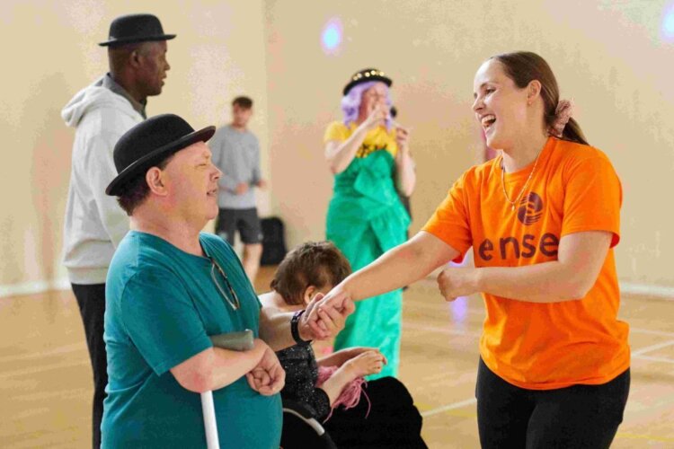 A male wearing a green T-shirt and a black bowler hat holding a walking aid, holds hands with a woman wearing an orange sense T-shirt. Both people are smiling and dancing amongst a crowd in the back ground